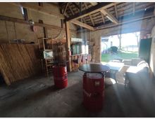 Ein rustikaler Innenraum mit offener Holzdachkonstruktion, zwei roten Fässern als Tische, einem Bereich mit Stühlen und Sofa sowie einem großen offenen Durchgang, der auf einen sonnigen Garten mit Rasen und Bäumen führt. Im Hintergrund sind weitere Möbel und Bambusmatten sichtbar.