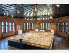 Ein Raum mit Backsteinwänden und spitzbogigen Fenstern, in dessen Mitte ein großes dreidimensionales Stadtmodell auf einem erhöhten Tisch präsentiert wird. Die Decke besteht aus dunklem Holz, und die Deckenbeleuchtung richtet sich auf das Modell.