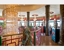 Chinesisches Restaurant mit aufwendiger Dekoration, darunter ein großer, funkelnder Kronleuchter, rote Gittergeländer, schwarze Marmorsäulen und farbenfrohe Vasen. Die Wände sind mit traditionellen chinesischen Mustern und grünen Fensterrahmen gestaltet.