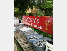 Ein Außenbereich mit einer roten Werbetafel mit der Aufschrift „Kant’s Kunst & Krempel“. Auf hölzernen Paletten stehen verzinkte Wannen, Kabel, Metallgegenstände und Kisten, umgeben von Bäumen und Vegetation.