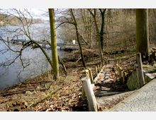 Ein Waldweg führt über eine steinerne Treppe mit hölzernen Geländern hinab zu einem See. Im Hintergrund ist ein schmaler Steg zu sehen, der ins Wasser ragt, umgeben von kahlen Bäumen.