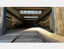 Leerer Industriehallenraum mit hohen Decken und großen Oberlichtern, die Tageslicht einlassen. Sichtbare Wände aus Beton und Ziegeln sowie eingebaute Metallrohre und Schienen im Boden prägen die Raumstruktur.