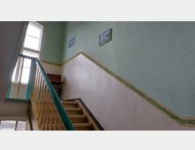 Treppenhaus in einem älteren Gebäude mit hellgrünen Wänden, beigefarbenen Stufen und einem hölzernen Geländer. An der Wand hängen kleine Bilder, und ein Fenster lässt Tageslicht in den Raum fallen.