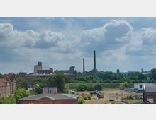 Industriegebiet mit alten Backsteingebäuden, grünen Bäumen und zwei hohen Schornsteinen im Hintergrund. Im Vordergrund befinden sich offene Flächen mit Baustellenfahrzeugen, und im Zentrum ein Gebäude mit der Aufschrift „RITAS“.