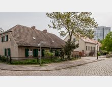 Straßenszene in einem malerischen Viertel mit Kopfsteinpflaster, einem traditionellen einstöckigen Haus mit Satteldach und grünen Fensterläden im Vordergrund. Im Hintergrund sind weitere Gebäude und ein moderner Wohnblock zu sehen, umgeben von Bäumen und Grünflächen.