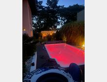 Ein beleuchteter Außenpool mit rotem Licht in einer Gartenanlage, umgeben von hohen Gräsern und Bäumen, mit einem modernen Gebäude und einer Terrasse im Hintergrund.