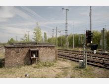 Backsteinbau in vernachlässigtem Zustand neben einem Bahngleis mit Oberleitungsmasten und Signalanlage im Hintergrund. Im Vordergrund befindet sich trockenes Gras, und ein kleines Bäumchen wächst aus dem Dach des Gebäudes.