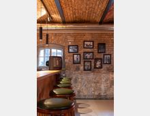 Ein rustikaler Barbereich mit Ziegelgewölbedecke, einer Holztresenfront und grünen Barhockern mit Polstern. Im Hintergrund sind eine Backsteinwand mit gerahmten Schwarz-Weiß-Bildern sowie ein Fenster und ein alter Röhrenfernseher zu sehen.