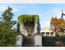 Steintreppe, die zu einer überdachten Pergola mit Weinranken führt, flankiert von zwei bronzenen Statuen; im Hintergrund ein klassizistisches Gebäude mit Säulen.
