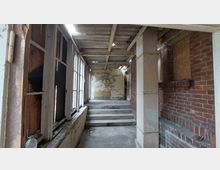 Verlassener Innenraum mit sichtbaren Ziegelwänden, abblätternder Putz an Wänden und Decken sowie kaputten Fenstern auf der linken Seite. Eine kleine Treppe führt zu einem erhöhten Bereich im Hintergrund.