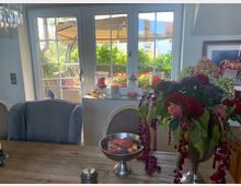 Ein geschmackvoll eingerichteter Innenraum mit einem Holztisch, dekorativen Stühlen und einem großen Blumenarrangement mit roten Rosen im Vordergrund. Im Hintergrund befindet sich ein Fenster mit Blick auf eine Terrasse mit Pflanzen, Gartenmöbeln und einem Sonnenschirm.