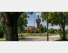Straßenszene bei einem Kreisverkehr mit Ampeln und einem Backstein-Wasserturm im Hintergrund, umgeben von Bäumen und einem Gebäude mit rotem Ziegeldach. Im Vordergrund fährt eine Person mit Fahrrad.