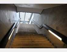 Treppenaufgang in einer U-Bahn-Station mit grauen Wänden, die mit Graffiti bedeckt sind, und Neonbeleuchtung an den Seiten. Oben ist ein Glasdach mit Metallstreben und Tageslicht erkennbar.