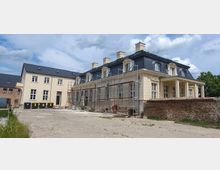 Historisches Gebäude mit gelblich-beigem Putz und mehreren Dachgauben, das von einem Gerüst für Renovierungsarbeiten umgeben ist. Daneben stehen ein modernes Nebengebäude und eine niedrige Ziegelmauer, die einen offenen Hof begrenzen.