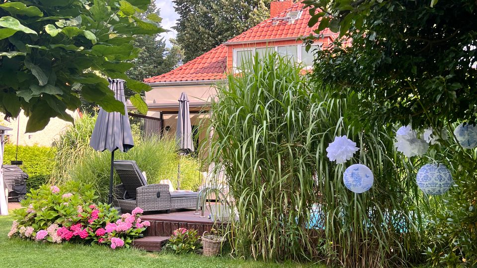 Blick in einen üppig bepflanzten Garten mit Rasenfläche, Holzterrasse und Sitzmöbeln samt Sonnenschirmen. Im Hintergrund steht ein Wohnhaus mit rotem Ziegeldach, umgeben von hohen Bäumen und dichtem Grün mit Blumenbeeten und hängenden Papierlaternen.