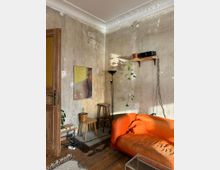 Ein Wohnzimmer mit einer abgenutzten Wand, einer orangefarbenen Couch, einem Holzboden und historisch verzierten Stuckdetails an der Decke. Ein Regal mit einer Pflanze und einem Lautsprecher, ein gerahmtes Bild sowie verschiedene kleine Möbelstücke wie Hocker und Tische schmücken den Raum.