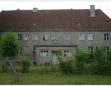 Ein zweistöckiges, verfallenes Gebäude mit einem steilen Ziegeldach und mehreren beschädigten Fenstern. Vor dem Gebäude stehen vereinzelt Bäume und Büsche, und ein einfacher Metallzaun trennt das Grundstück von einer Grasfläche im Vordergrund.