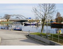 Hafenbereich mit mehreren festgemachten Booten und einer modernen Brücke mit geschwungenem Stahlbogen im Hintergrund. Im Vordergrund Gehwege, Geländer und ein Baum, umgeben von Grünflächen.