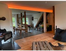 Wohnraum mit offener Küche in rustikalem Stil, geprägt von Holzbalken und Holzboden. Der Raum umfasst einen Esstisch, offene Regale, eine Treppe und eine moderne Feuerstelle; große Fenster lassen Tageslicht herein.