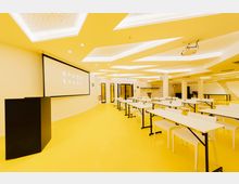 Ein moderner Seminarraum mit gelbem Boden, weißen Tischen und Stühlen, einer großen Projektionsfläche mit der Aufschrift „Space Shack Coworking" sowie einer Decke mit geometrischen LED-Leuchtelementen. Im Hintergrund befinden sich Glastüren und eine Garderobe.