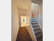 Ein schmaler Flur mit weiß gestrichenen Wänden und einer Treppe mit Holzstufen, die rechts nach oben führt. Der Boden des Flurs besteht aus rustikalen Ziegeln, und im Hintergrund ist ein Raum mit hellem Holzboden zu erkennen.