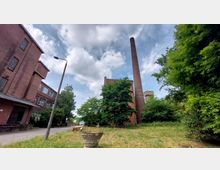 Industriegelände mit roten Backsteingebäuden, einem hohen Schornstein und umliegendem Grünbereich mit Bäumen und Gras. Im Vordergrund befindet sich eine Wiese mit einem Betonpflanzgefäß, und ein Teil einer Straße ist sichtbar.