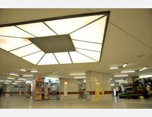 Unterirdisches Einkaufszentrum mit hell beleuchteter Decke in geometrischem Design. Links ein Kiosk mit Zeitungen und Plakaten, rechts ein Blumenladen mit Topfpflanzen, umgeben von gefliesten Wänden und Hinweisschildern.