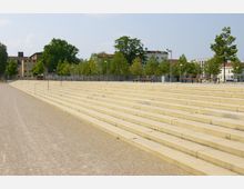 Weitläufige Treppenanlage aus beigem Stein, die zu einer Baumreihe mit Sitzbänken und Laternen führt, im Hintergrund sind mehrstöckige Gebäude und große Bäume zu sehen.