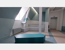 Heller Raum mit modernem Design, geprägt von schrägen Wänden und einer hellblau-weißen Farbgestaltung. Sichtbare Elemente sind ein Heizkörper, ein Fenster, geometrisch geformte Sitzmöbel und eine durchgehende Linienführung, mit einer zentralen Säule und angedeuteten Podesten.