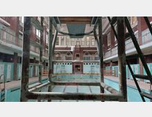 Historisches Hallenbad mit leerem Schwimmbecken, hohen Decken und großen Fenstern. Die Wände sind mit türkisfarbenen Kacheln verkleidet, und es gibt Umkleidekabinen sowie eine verrostete Sprungturmkonstruktion im Vordergrund.