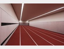 Ein moderner Innenraum mit einer roten Laufbahn, bestehend aus mehreren weißen Bahnen, flankiert von hellen Akustikwänden. Die Decke ist mit linearen Leuchtstofflampen beleuchtet.