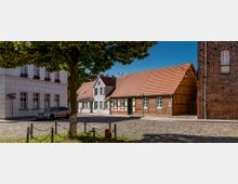 Straßenszene in einer historischen Altstadt mit Kopfsteinpflaster. Links ein weißes Gebäude mit Blumen in den Fensterkästen, in der Mitte zwei Fachwerkhäuser mit roten Ziegeldächern, und rechts ist ein gemauertes Gebäude sichtbar. Im Vordergrund steht ein Baum.