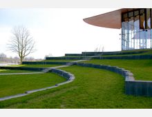 Geschwungenes, terrassenförmiges Gelände mit grünen Rasenflächen und gepflasterten Wegen, die durch niedrige Betonmauern getrennt sind. Rechts im Bild ist ein modernes Gebäude mit einer Glasfassade und einem breiten, geschwungenen Dach zu sehen, im Hintergrund ein alleinstehender Baum und ein Wasserbereich.
