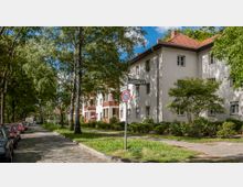 Straßenszene in einer Wohngegend mit gepflastertem Gehweg und parkenden Autos auf der linken Straßenseite. Rechts sind mehrstöckige Wohnhäuser mit hellen Fassaden und roten Dächern zu sehen, umgeben von Bäumen und gepflegten Grünflächen.