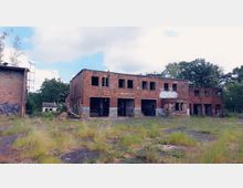 Verlassene Backsteingebäude mit eingeschlagenen Fenstern und Graffiti, umgeben von überwuchertem Gelände mit Gras und Bäumen im Hintergrund. Die Gebäude haben große Toröffnungen, die einst industriellen oder gewerblichen Zwecken dienten.