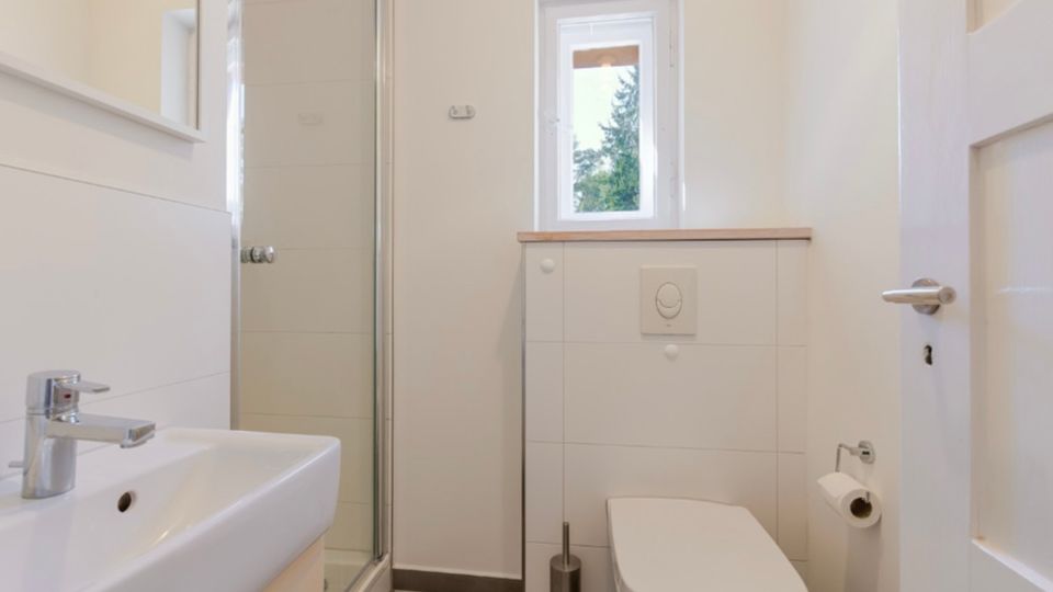Kleines, helles Badezimmer mit weiß gefliesten Wänden, Waschbecken im Vordergrund, einer Glasduschkabine und einer wandhängenden Toilette. Über dem Spülkasten befindet sich ein schmales Fenster; der Boden ist dunkel gefliest.