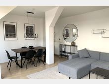 Modernes Wohnzimmer mit Essbereich. Ein Holztisch mit schwarzen Stühlen steht unter einer Hängelampe, daneben ein graues Ecksofa, ein runder Wandspiegel und ein minimalistisches Metallregal auf hellem Holzboden.