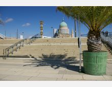 Steintreppe mit Metallgeländern führt zu einer Kuppelkirche im Hintergrund, umgeben von Baugerüsten. Im Vordergrund steht ein großer Palmenbaum in einem grünen Pflanzkübel, daneben eine verzierte Steinsäule.