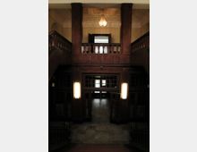 Holzverkleidetes Foyer eines historischen Gebäudes mit einer zentralen Treppe, flankiert von Geländern aus Holz. Im oberen Bereich befindet sich ein offener Balustradenabschnitt, von dem ein Kronleuchter herabhängt. Türen mit Glaseinsätzen und Wandlampen prägen den Eingangsbereich.