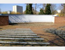 Freiluftbühne mit halbrunder, gepflasterter Spielfläche und einer weißen Rückwand, umgeben von roten Ziegelmauern. Mehrere Reihen einfacher, metallener Sitzbänke auf einem gepflasterten Platz; im Hintergrund sind Bäume und moderne Gebäude zu sehen.