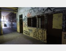 Ein verlassenes, heruntergekommenes Gebäude mit beschädigten Wänden, die mit Graffiti bedeckt sind. Die Szene zeigt einen dunklen Flur mit offenen Räumen, zerbrochenen Fenstern und freiliegenden Rohren an der Decke.