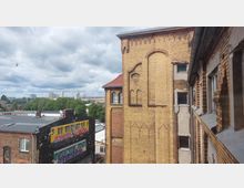 Backsteinfassade eines historischen Gebäudes mit verzierten Fenstern und abgestuften Mauerwerkdetails im Vordergrund. Im Hintergrund sind ein modernes Wandgemälde auf einem Gebäude, Bäume sowie Hochhäuser unter wolkigem Himmel sichtbar.