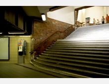 U-Bahnhof-Treppenaufgang mit beige gefliesten Wänden und einer gemauerten Seitenwand, links ein öffentlicher Münztelefonautomat, rechts führen breite Steinstufen nach oben zu einem beleuchteten Ausgang.