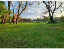 Weitläufige Wiese mit vereinzelten, blattlosen und belaubten Bäumen, im Hintergrund Ruinen einer historischen Backsteinstruktur sowie weitere Gebäude. Die Szenerie ist von grünem Gras und einer bewaldeten Umgebung geprägt, mit einem teils bewölkten Himmel.