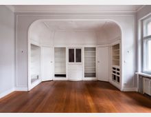 Leerer Innenraum mit hoher Decke und großem, bogenförmigem Wandeinbau aus weißen Einbauschränken und Regalen, inklusive Schubladen in der Mitte. Davor liegt ein dunkler Holzdielenboden; rechts befindet sich ein großes Fenster mit Heizkörper darunter.