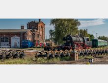 Stillgelegtes Eisenbahngelände mit einer historischen Dampflok, mehreren Waggons und gelagerten Eisenbahnrädern auf einem Gleisbett. Im Hintergrund sind Backsteingebäude mit industriellem Charakter und Bäume zu sehen.