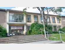 Zweistöckiges Gebäude mit beigen Fassaden, großen Fenstern und einem Eingang mit Glastüren, über dem ein Schild mit der Aufschrift „Stasi-Zentrale. Campus für Demokratie“ angebracht ist. Vor dem Eingang führen Stufen, umgeben von Büschen und einem Baum, zum Gebäude hinauf.