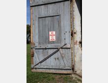 Große, verwitterte Holztür in hellem Grau mit diagonalen Verstrebungen, teils offenstehend, an einem alten Gebäude mit teilweise freiliegendem Backstein. Ein Schild mit der Aufschrift „No Parking Any Time“ ist in der Mitte der Tür angebracht.
