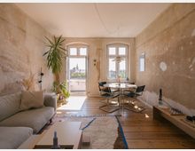 Wohnzimmer im minimalistischen Stil mit rauen, unbehandelten Wänden und Holzdielenboden. Im Vordergrund ein beiges Sofa, ein niedriger Couchtisch und eine Zimmerpflanze, im Hintergrund ein runder Esstisch mit vier Stühlen vor doppelflügeligen Fenstertüren, die auf einen Balkon führen und den Blick auf eine Stadtlandschaft mit Kirchturm freigeben.