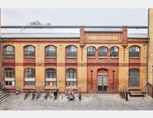 Historische Backsteinfassade eines zweistöckigen Gebäudes mit großen Fenstern und Torbogen-Elementen, umgeben von einem gepflasterten Innenhof. Im Vordergrund stehen mehrere Fahrräder, eine Holzbank und ein Müllbehälter.